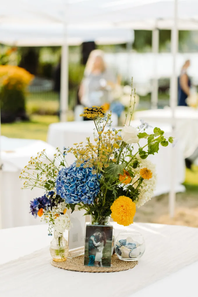 Blue hydrangea and marigold wildflower centerpiece with a framed engagement photo on a linen-covered table