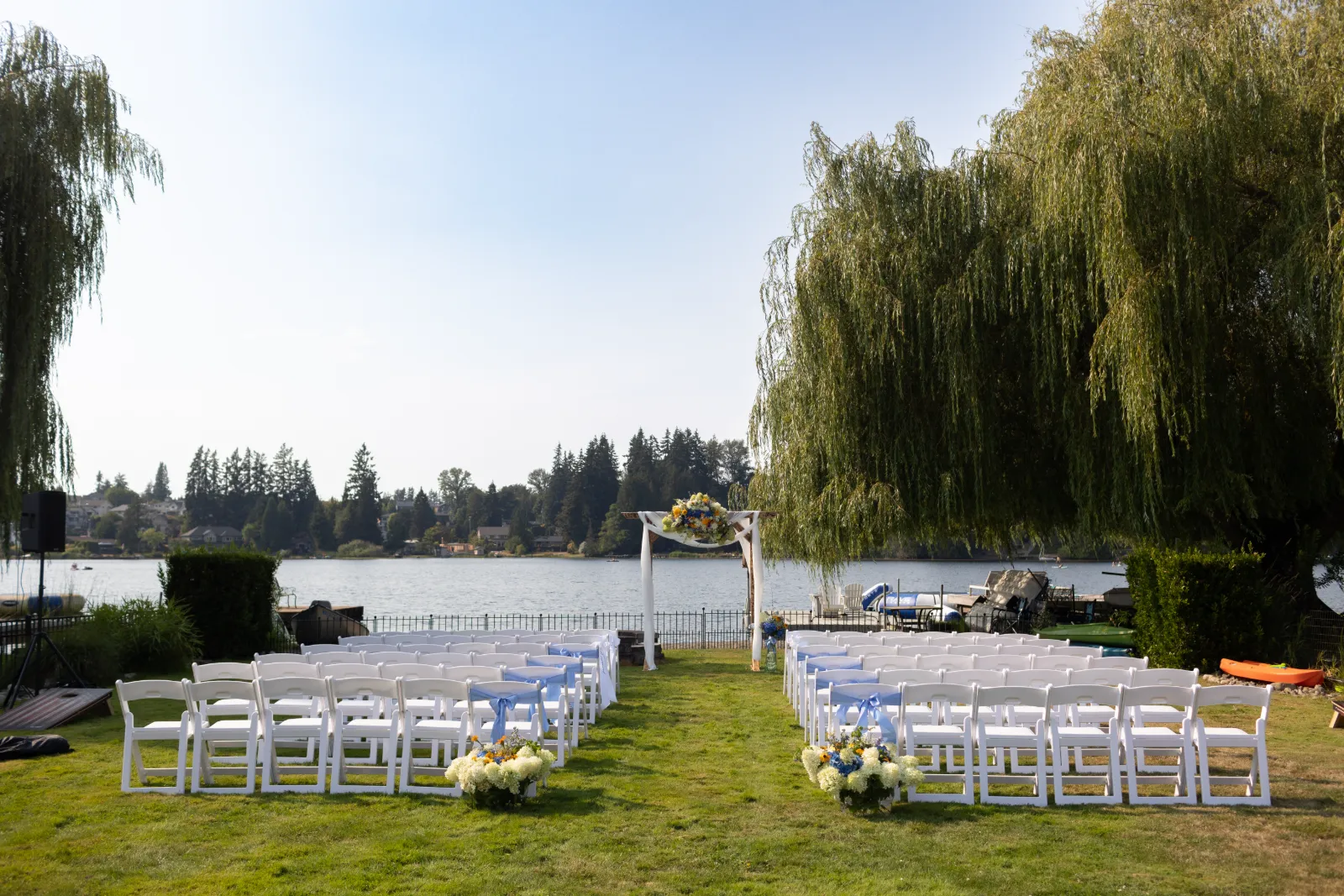 Lakeside ceremony setup with white chairs, blue sashes, and a floral arch beneath a willow tree