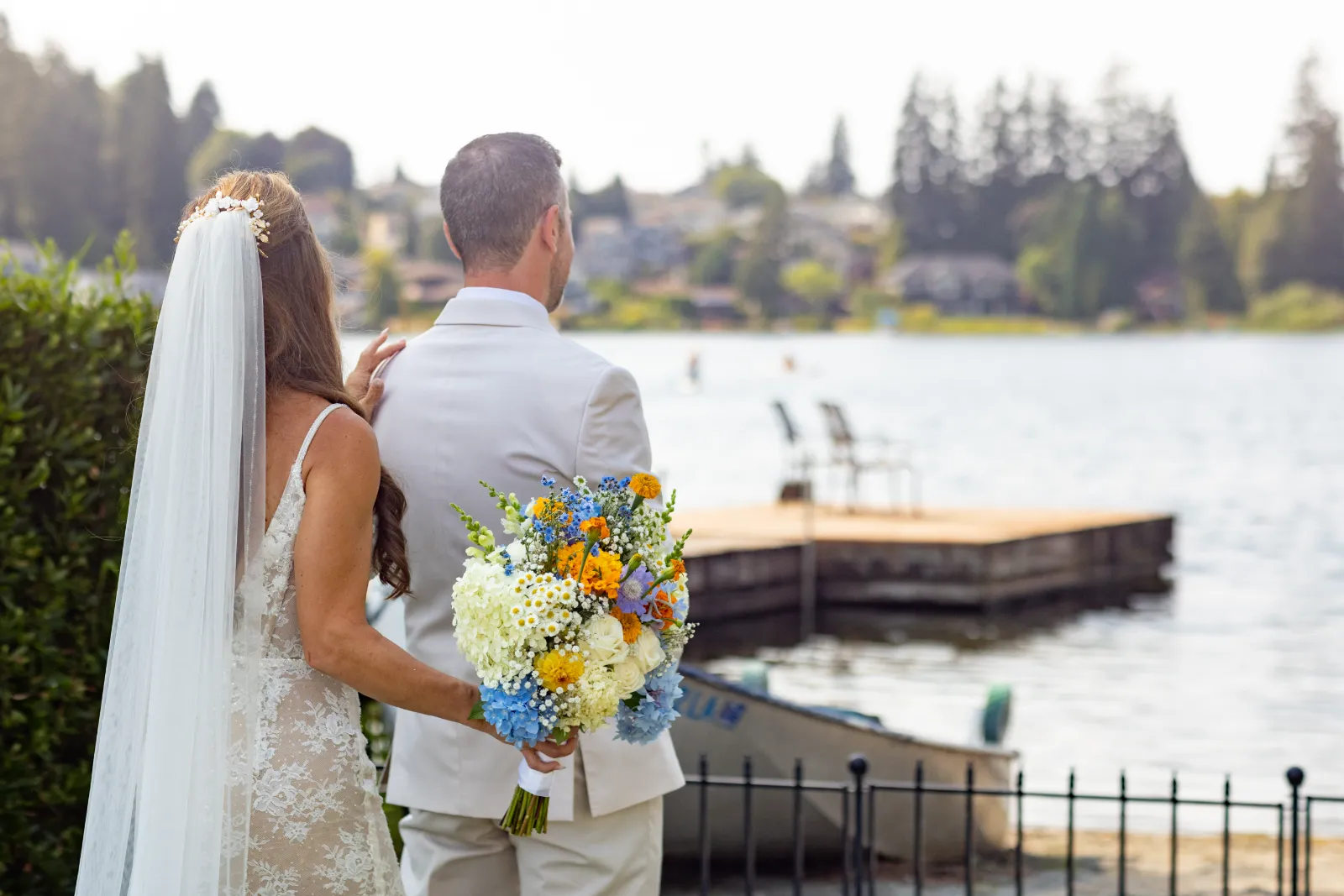 Bride touching the groom's shoulder from behind during a first look by the lake, holding a blue and yellow bouquet
