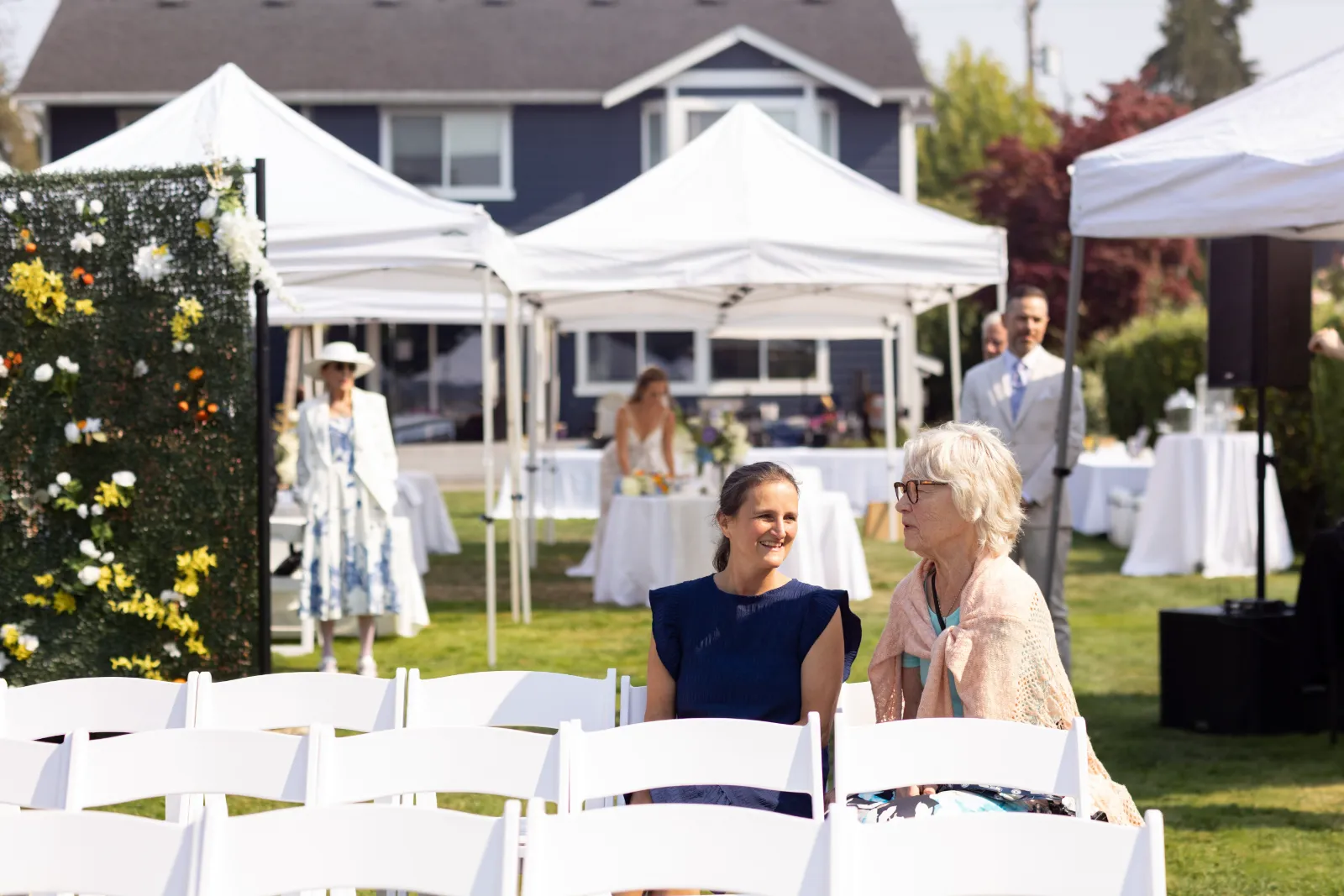 Two guests chatting in white chairs before the ceremony, with white tents and a floral backdrop behind them