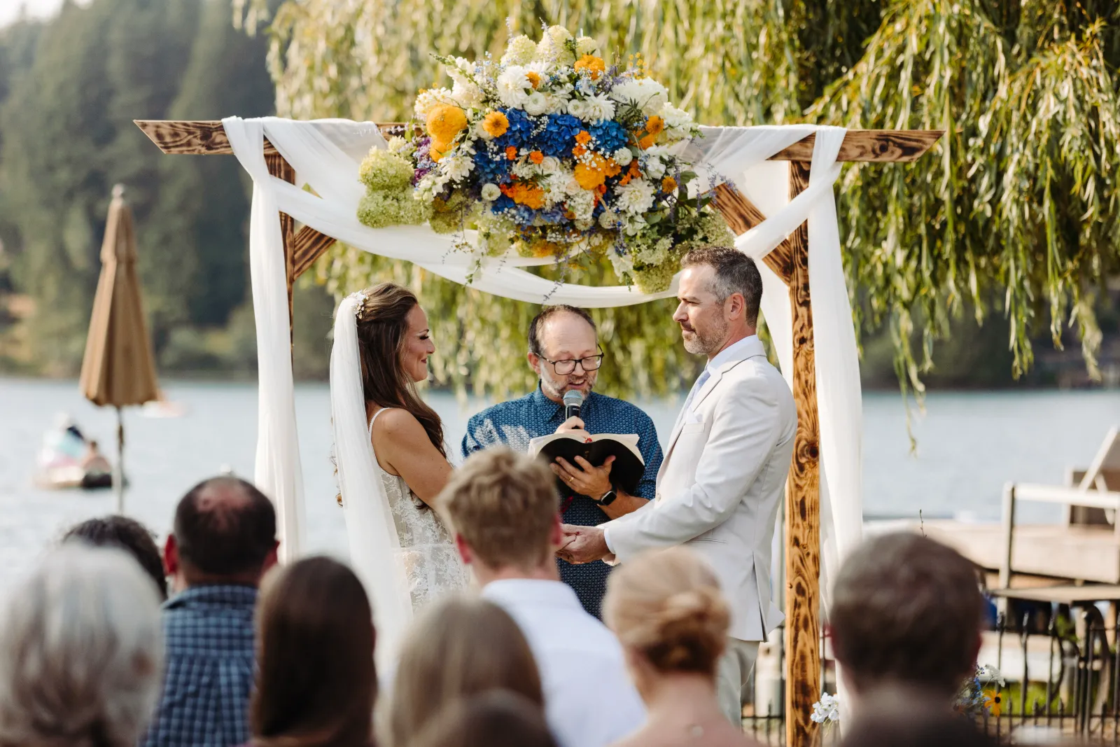 Bride and groom holding hands under a wooden arch with a large blue, white, and yellow floral piece as the officiant reads