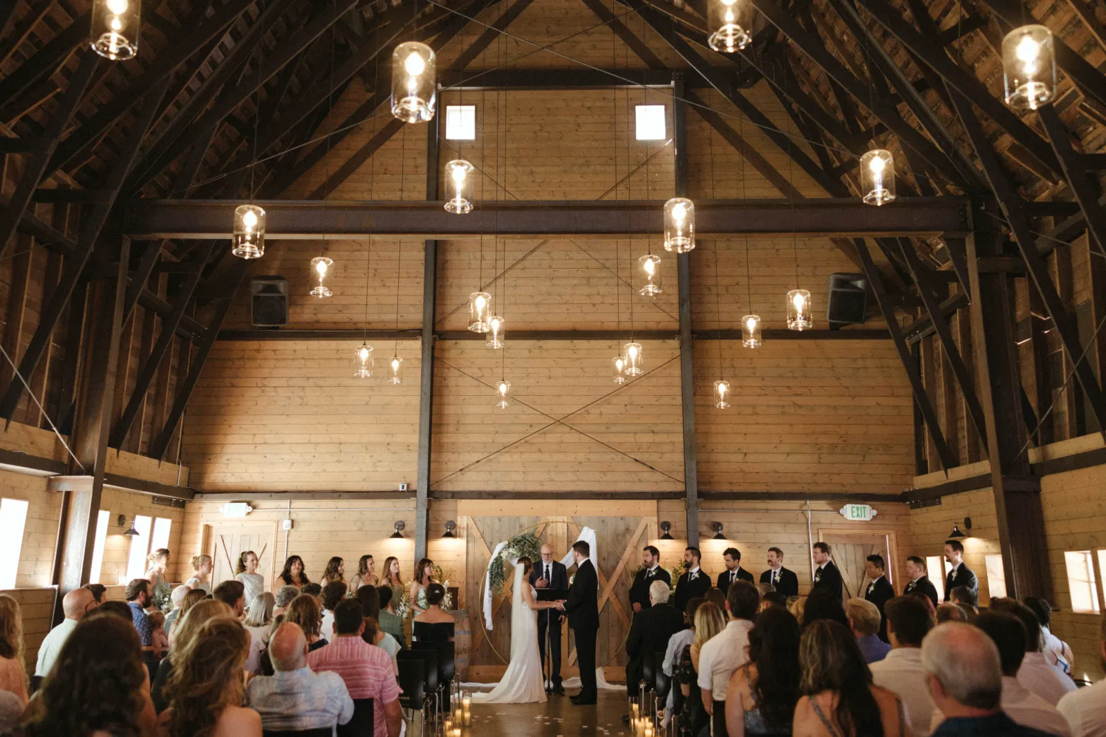 Wedding ceremony inside a timber-frame barn with hanging pendant lights, the couple at a round gold arch