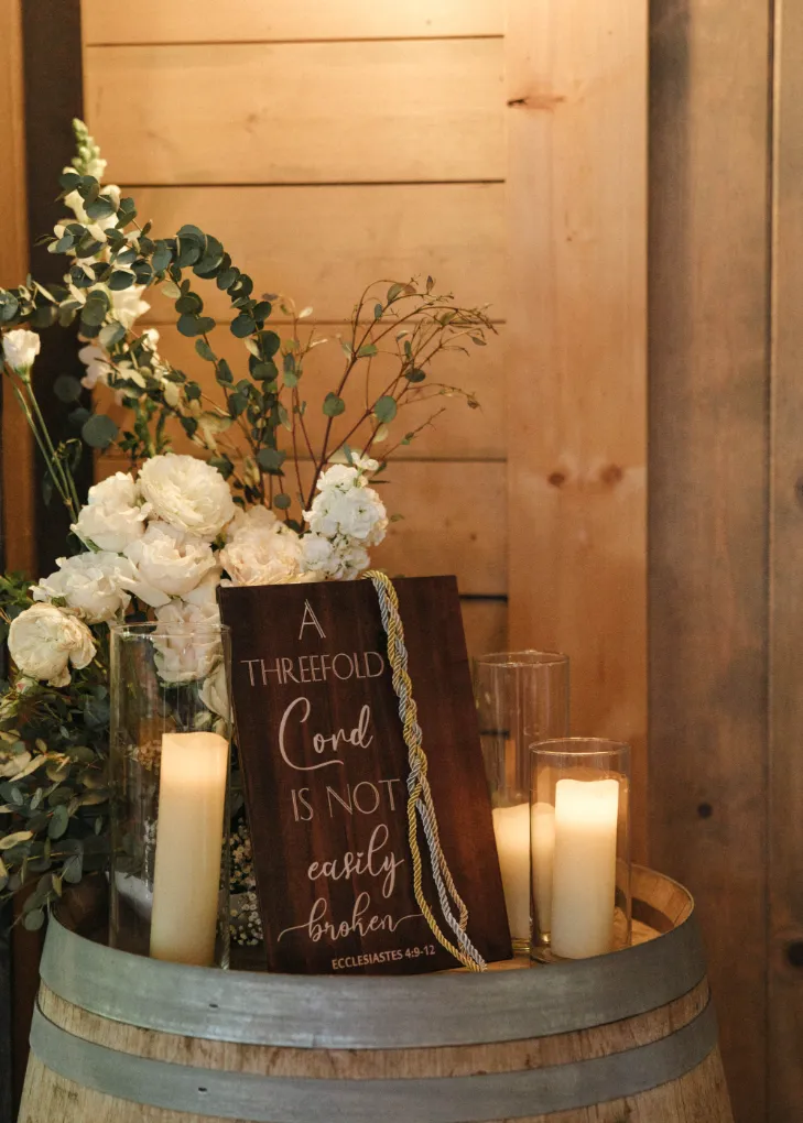 Wooden A Threefold Cord Is Not Easily Broken sign with a braided cord, candles, and white flowers on a wine barrel
