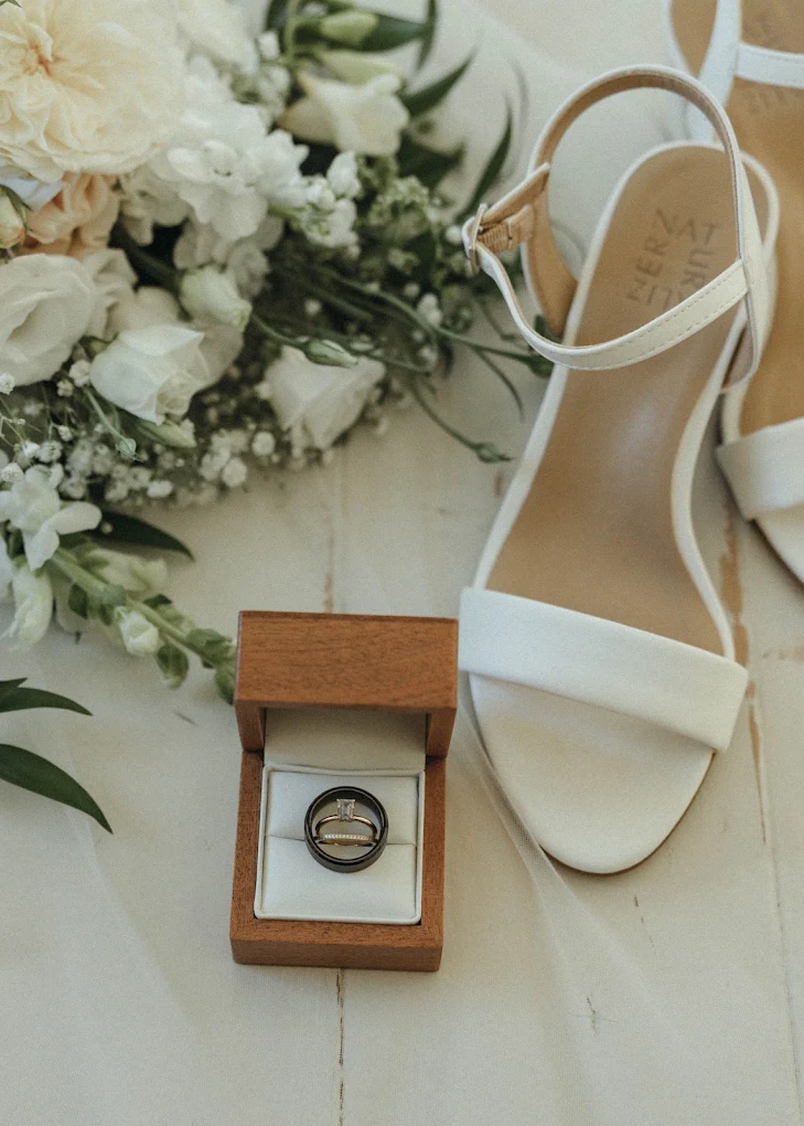 Bridal bouquet, white heels, and wedding rings displayed in a wooden box
