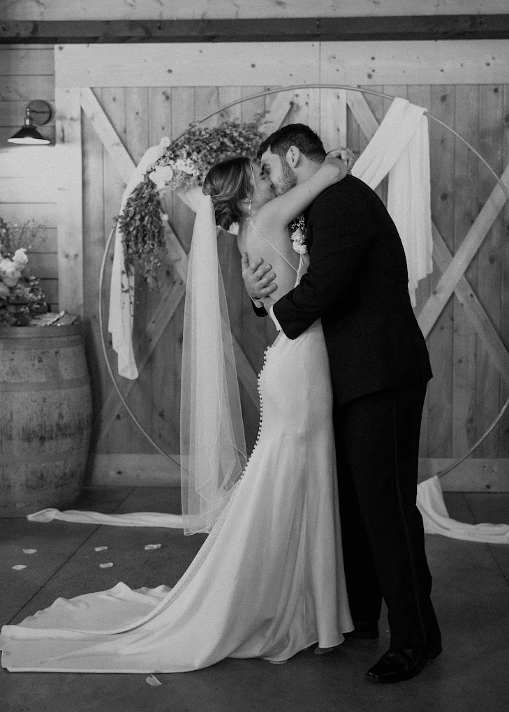 Black-and-white photo of bride and groom kissing at the altar inside a barn venue