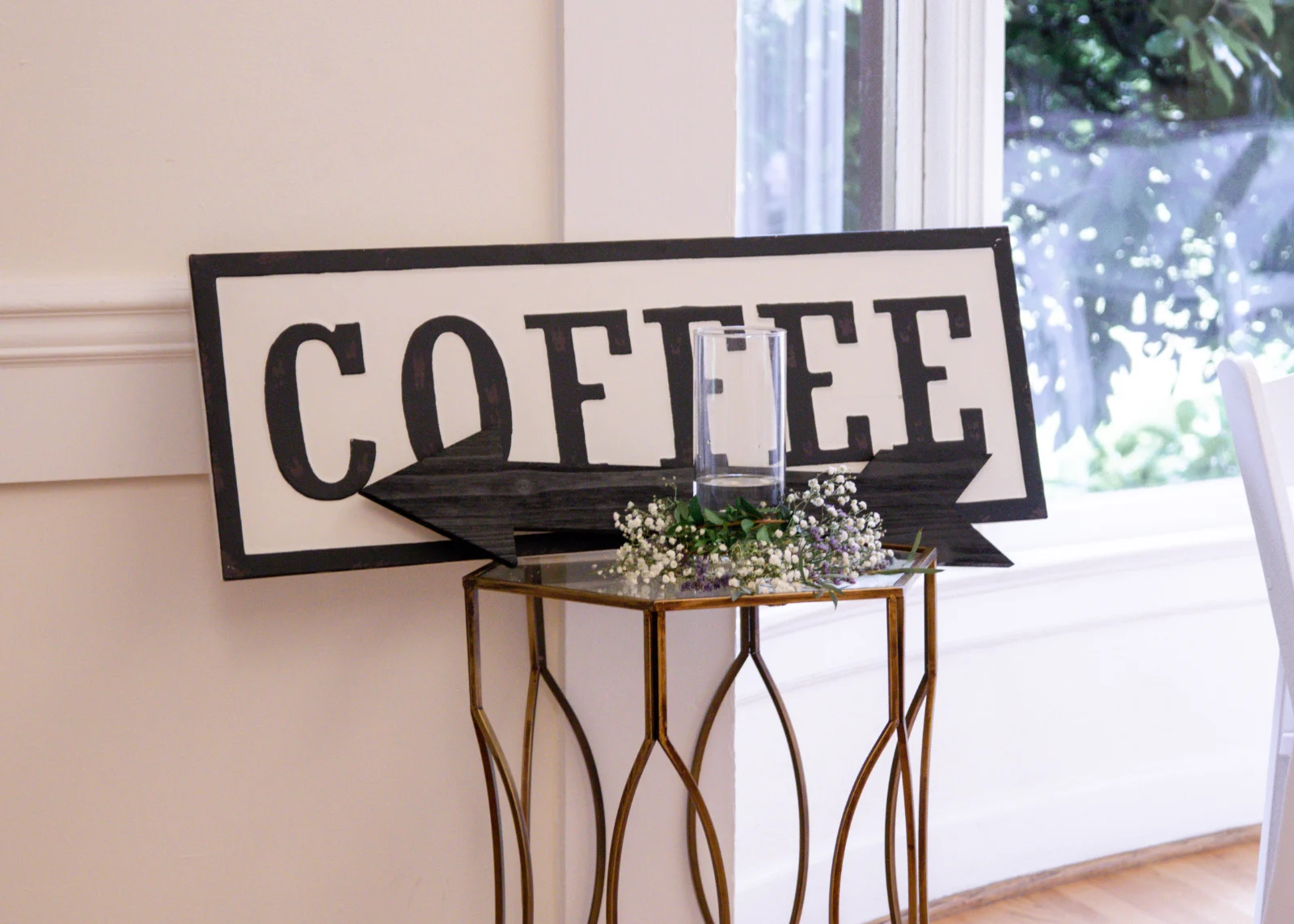 Black-and-white COFFEE sign with an arrow on a gold side table with baby's breath, beside a sunny window