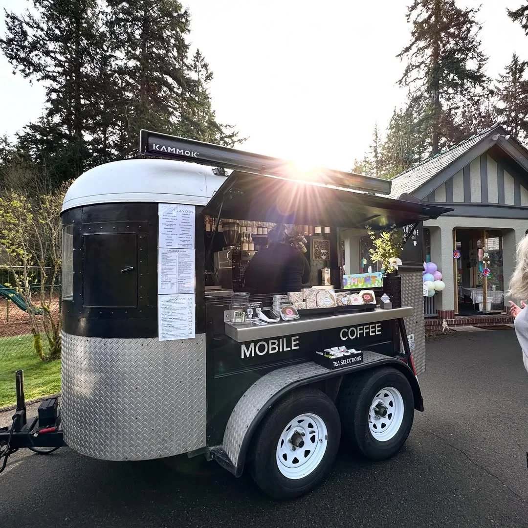 Mobile coffee trailer with menu and pastries on display, parked beside a park cottage with pastel balloons